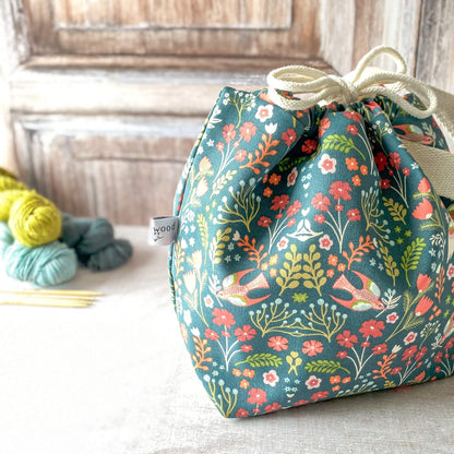 A close-up of a medium-sized knitting project bag featuring colorful birds and flowers in red, orange, and teal on a dark green background. The bag has sturdy fabric handles and a drawstring closure. Skeins of yarn and knitting needles are visible in the background.