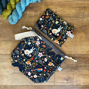  A knitting project bag and notions pouch sit flat on the floor. Both the bags are made using a black Scandi inspired folk art print. 