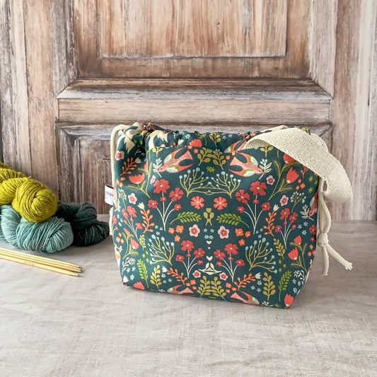 A small, drawstring knitting project bag featuring a vibrant floral and bird design in shades of red, orange, and teal against a dark green background. The bag has sturdy fabric handles and is placed on a textured surface with skeins of yarn and knitting needles nearby.