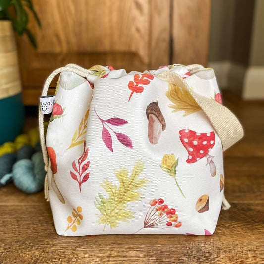 A project bag sits on a wooden floor. It is made from an autumnal print fabric featuring toadstools, acorns, berries and flowers all on a white background. Behind the bag can be seen three skeins of yarn and a houseplant. 
