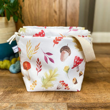 Load image into Gallery viewer, An open project bag sits on a wooden floor. It is made from an autumnal print fabric featuring toadstools, acorns, berries and flowers all on a white background. Behind the bag can be seen three skeins of yarn and a houseplant.