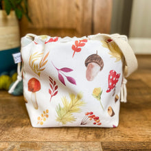 Load image into Gallery viewer, A close up image of an autumnal knitting and crochet project bag. The bag is made from fabric that shows off classic autumnal elements such as toadstools and acorns. 