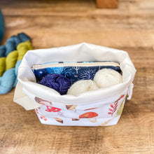 Load image into Gallery viewer, A project bag sits open on a wooden floor allowing the user to see inside. It holds three balls of yarn, one blue and two white, as well as a zipped pouch holding notions. Three skeins of yarn can also be seen on the floor behind the project bag. 