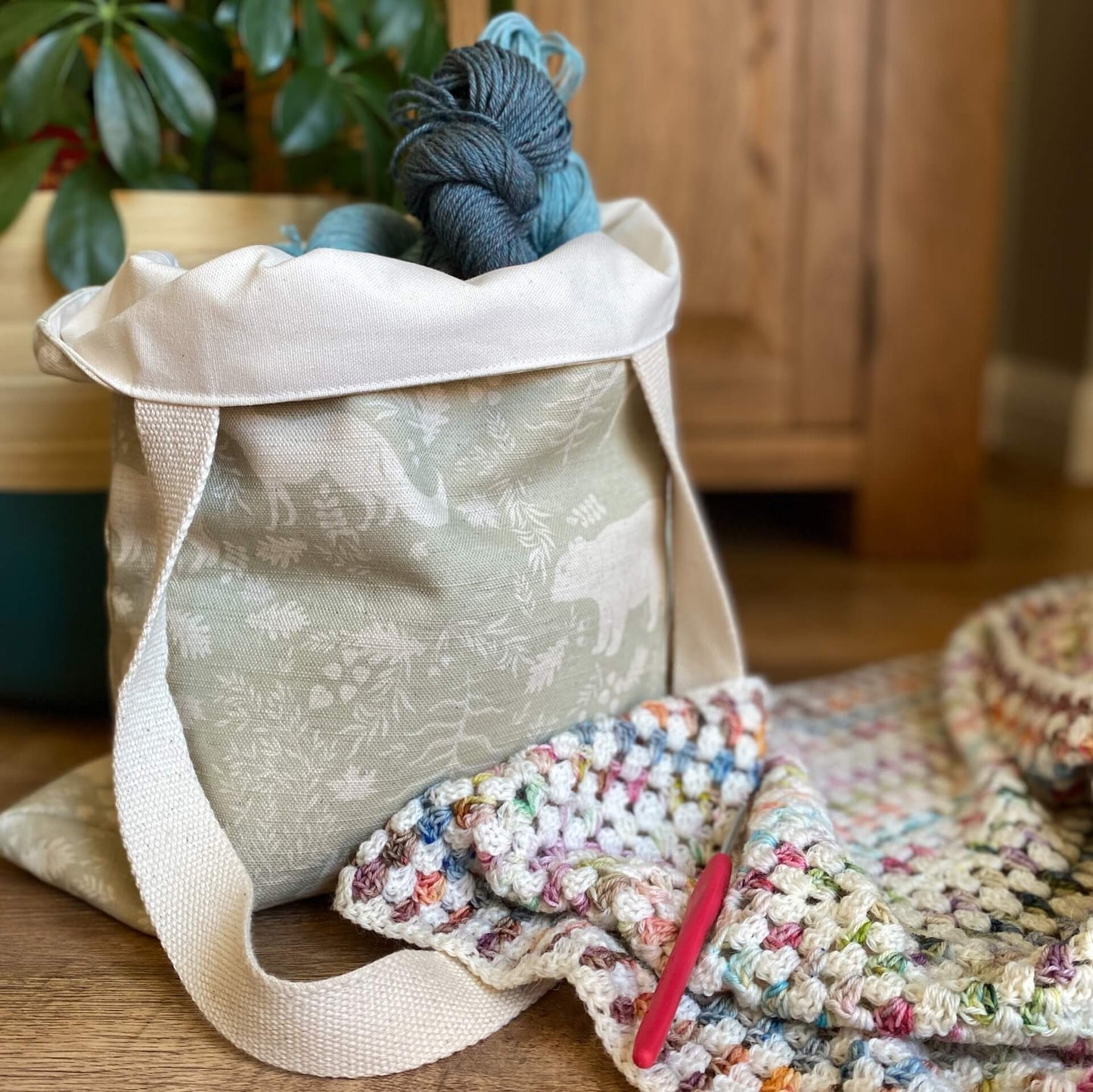A tote bag holding yarn sits on the floor next to a crochet blanket and a houseplant. The bag is made from a sage coloured fabric that is printed with woodland images. A crochet hook is sitting on the crochet blanket. 