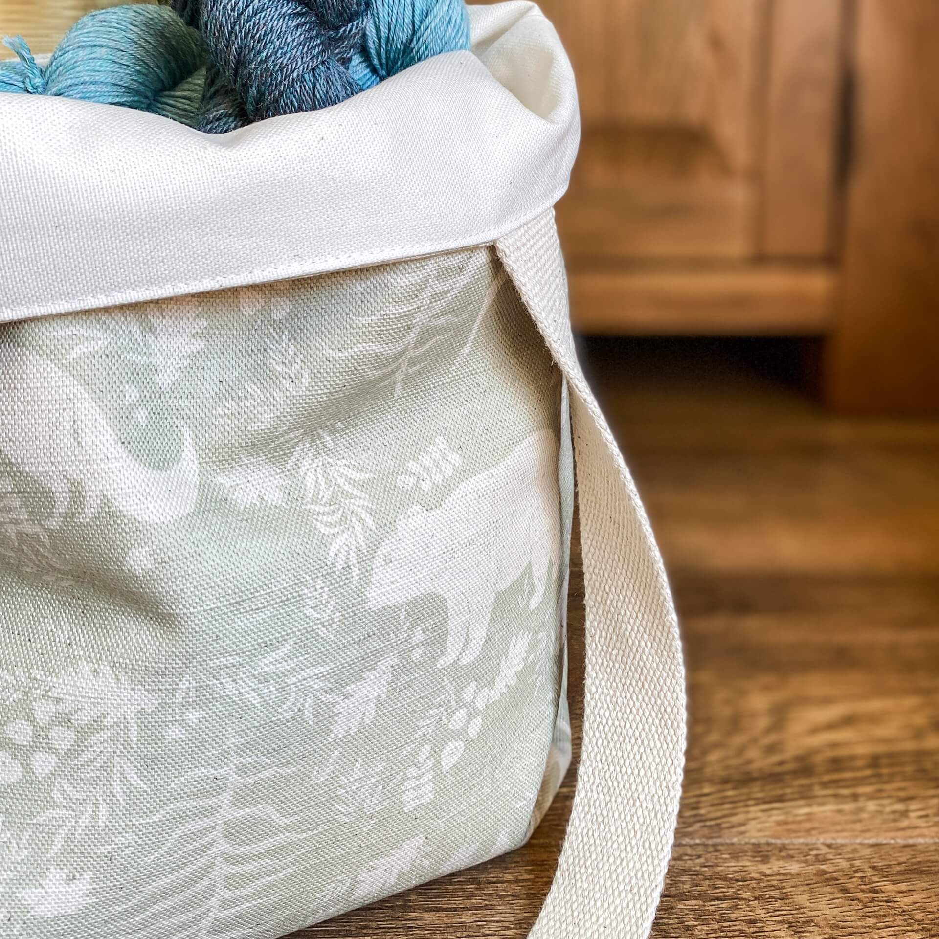 A close up image of a tote bag holding some blue yarn. The bag is made from a sage green fabric that is showing woodland animals and flora. The bag is sitting on a wooden floor.