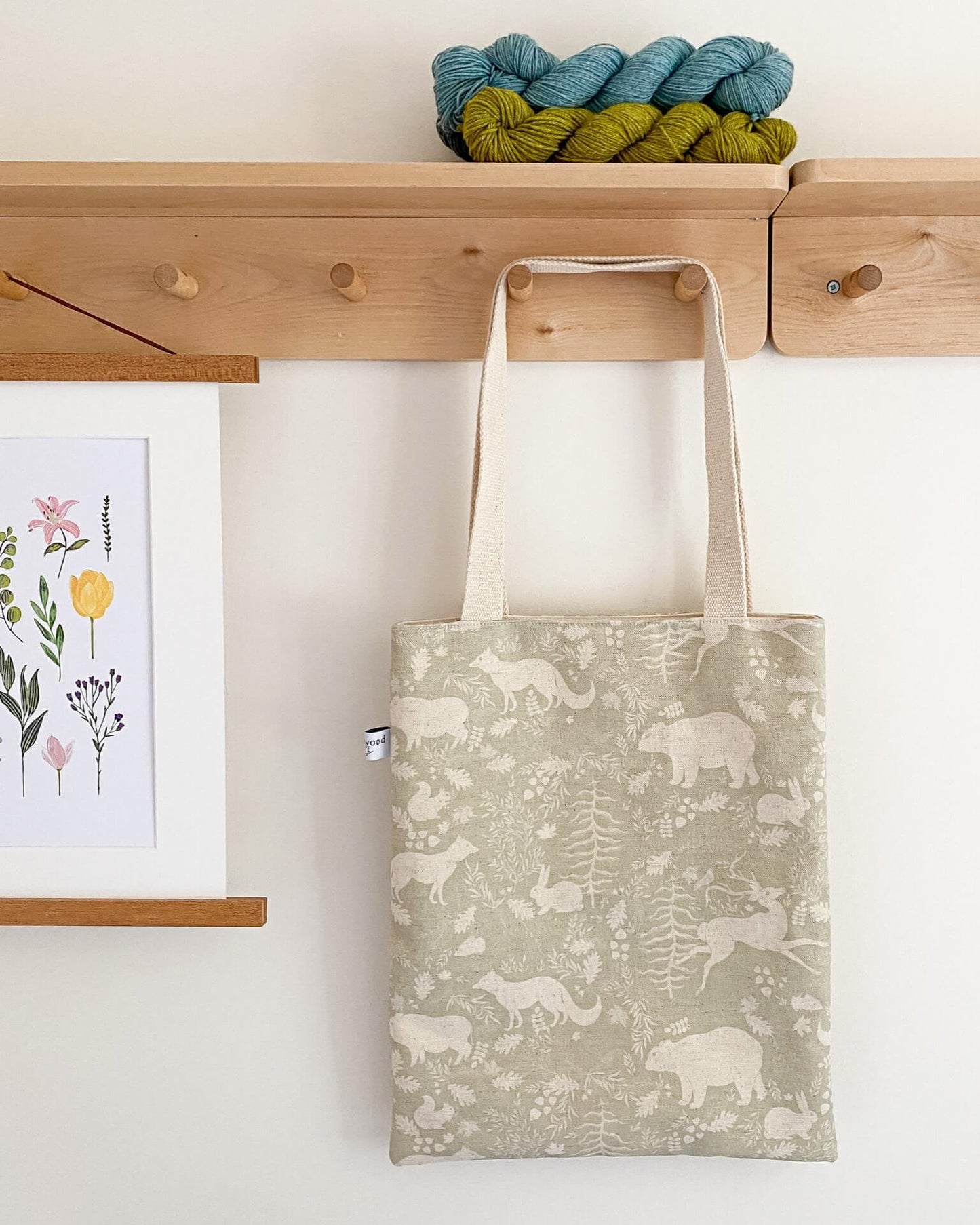 A sage green tote bag hangs from a set of wooden hooks. The bag is sage green in colour and covered in a woodland print. It has two long cream coloured handles. Above the bag on a shelf are some hanks of yarn and hanging next to the bag is a floral print.