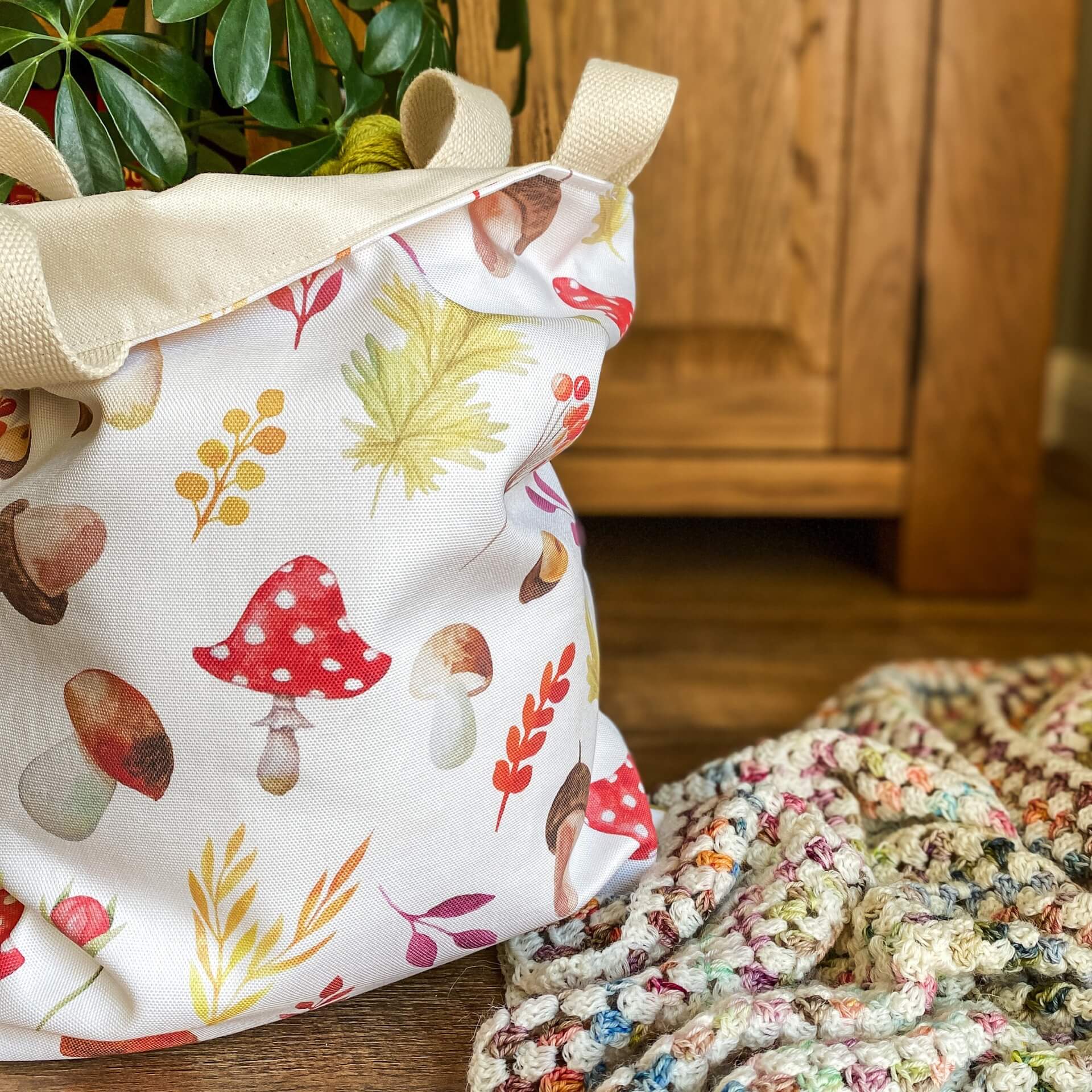 An autumnal tote bag holding yarn is on the floor next to a crochet blanket and a house plant. The bag is made from a white fabric that has lots of colourful images from an autumnal woodland printed on it. 