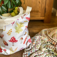 Load image into Gallery viewer, A tote bag filled with yarn sits next to a crochet blanket and a houseplant. The bag is made from an autumnal print with toadstools, acorns and leaves on it. 