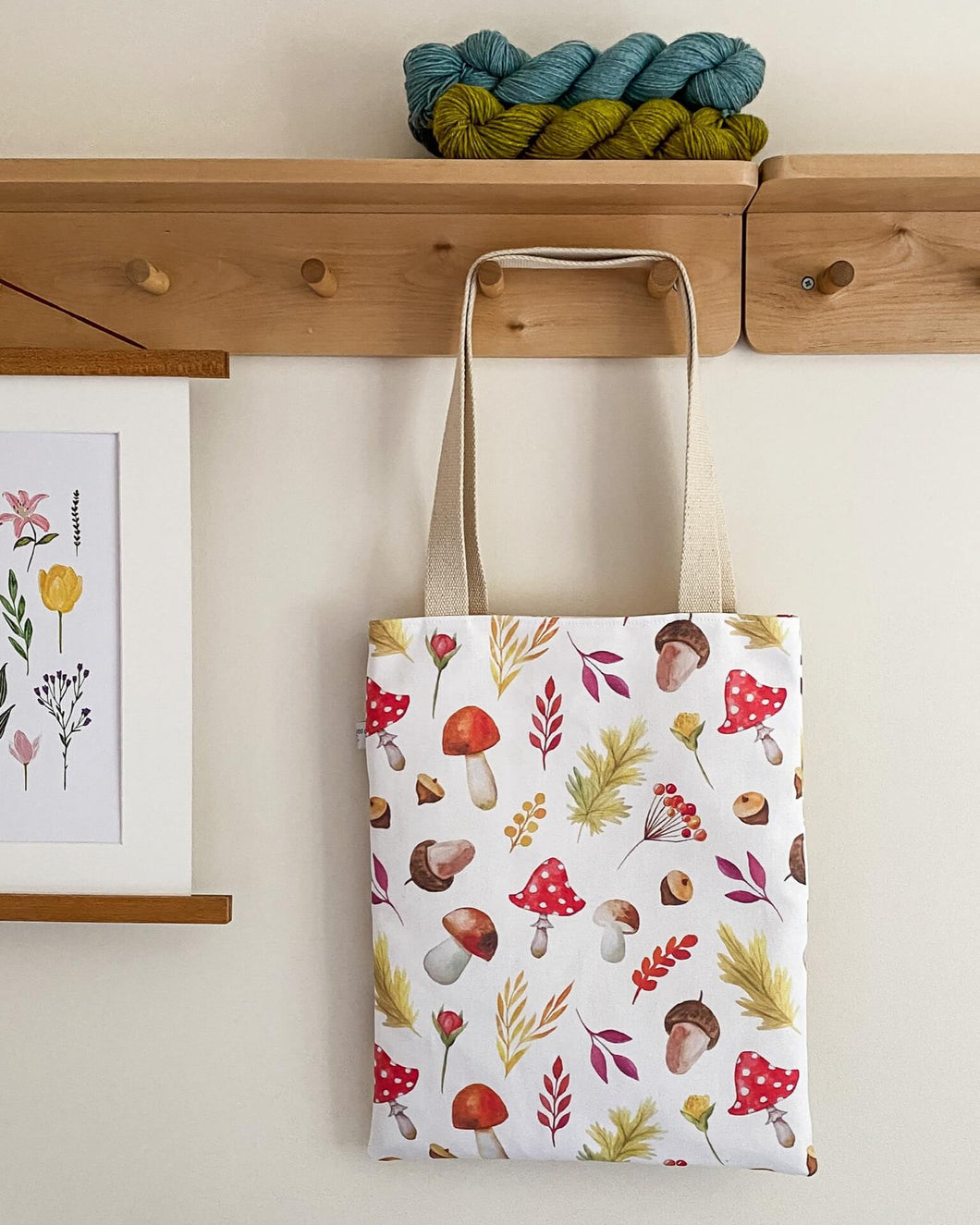 A tote bag hangs from a set of wooden hooks. The bag is covered in autumnal motifs such as toadstools and acorns. It has two long cream coloured handles. Above the bag on a shelf are some hanks of yarn and hanging next to the bag is a floral print.