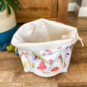 An empty project bag sits on the floor next to skeins of yarn. The bag is folded down so we can see the empty inside. 