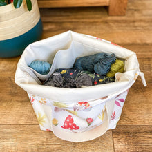 Load image into Gallery viewer, Looking into a knitting project bag which holds lots of yarn and another project bag. The main bag is sitting on the floor next to a plant pot and we can just see the leaves of the plant coming in at the edge. The project bag has lots of autumnal images printed on it like acorns and toadstools. 