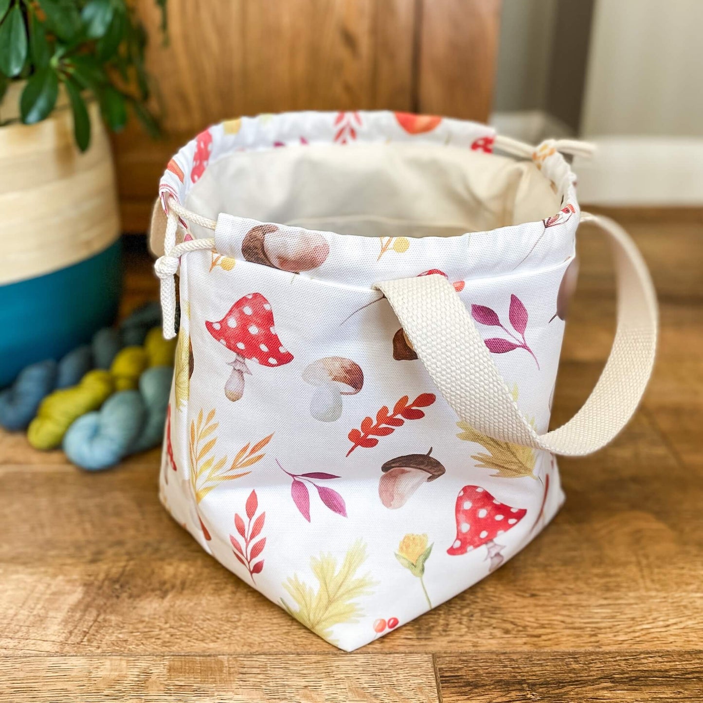 A large drawstring project bag is sitting side on to the camera and open wide. It shows that the inside is made from a creamy coloured canvas and the outside has lots of autumnal imagery on it such as toadstools and acorns. Next to the bag are three hanks of yarn and a house plant. 