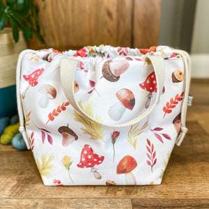 A large autumnal drawstring bag designed for craft projects is on the floor, pulled closed. The bag sits in front of some skeined up yarn and a large houseplant. 