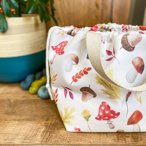 Part of a large drawstring bag is on the right hand side of the image. The bag is made from autumnal fabric showing mushrooms and toadstools and autumn leaves. There are three skeins of yarn on the floor next to the bag and a large house plant.
