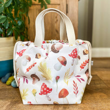 Load image into Gallery viewer, A large project bag made from autumnal woodland fabric is on a wooden floor. It has its drawstrings pulled closed and two cream handles sit proudly upright. Behind the bag, also on the floor, are three skeins of yarn and a houseplant. 