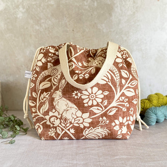 A large sized handmade project bag in a copper coloured heathland themed fabric. A drawstring closure at the top, and a sturdy handle, ideal for carrying knitting or crochet supplies and projects. Ideal for organising large knitting and crochet projects such as garments and blankets. The interior of the bag is made from a cream canvas and has lots of storage pockets. The bag sits in front of three skeins of yarn and a pot plant. 