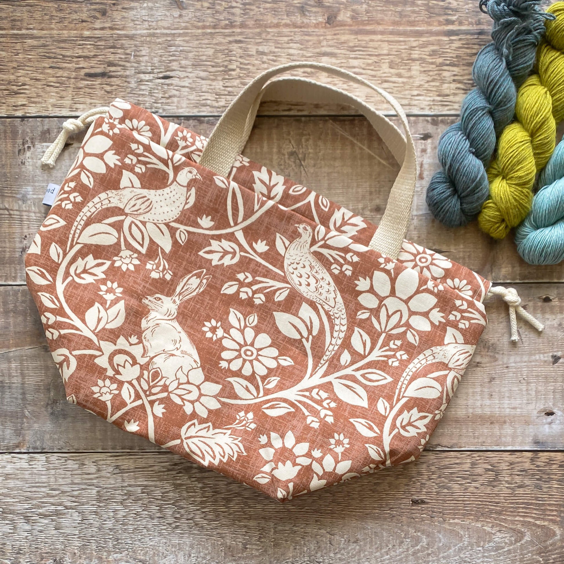 A large sized handmade project bag in a copper coloured heathland themed fabric, featuring a drawstring closure and sturdy handle, perfect for carrying knitting or crochet supplies. The bag is displayed next to three skeins of yarn and some wooden knitting needles. 