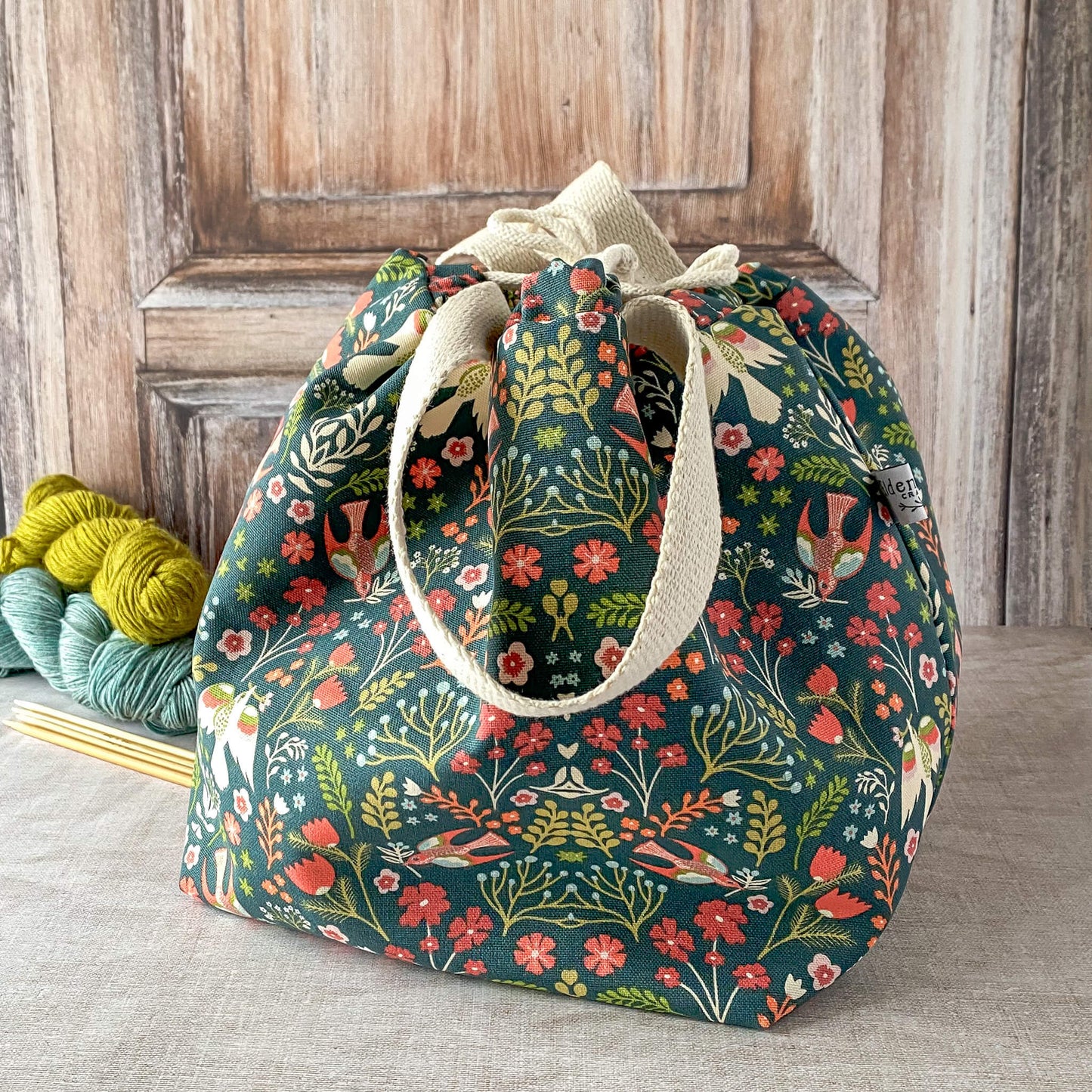 This image shows a large knitting project bag with a vibrant floral design featuring flowers and birds in shades of red, orange, and teal against a dark green background. The bag is cinched at the top with a drawstring, and its handles are made of light-colored fabric. It is placed on a neutral surface with skeins of yarn and knitting needles visible in the background.