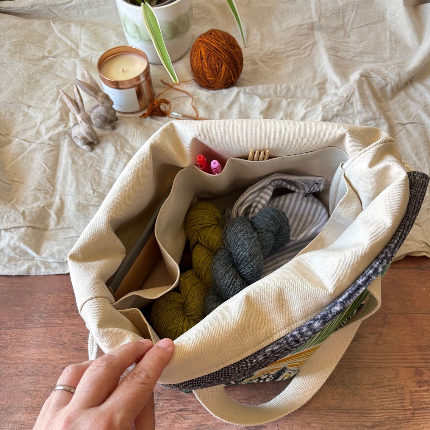 An open canvas tote bag with interior pockets, shown on a wooden surface with yarn skeins, a candle, and other craft items inside. A hand is holding the bag open.