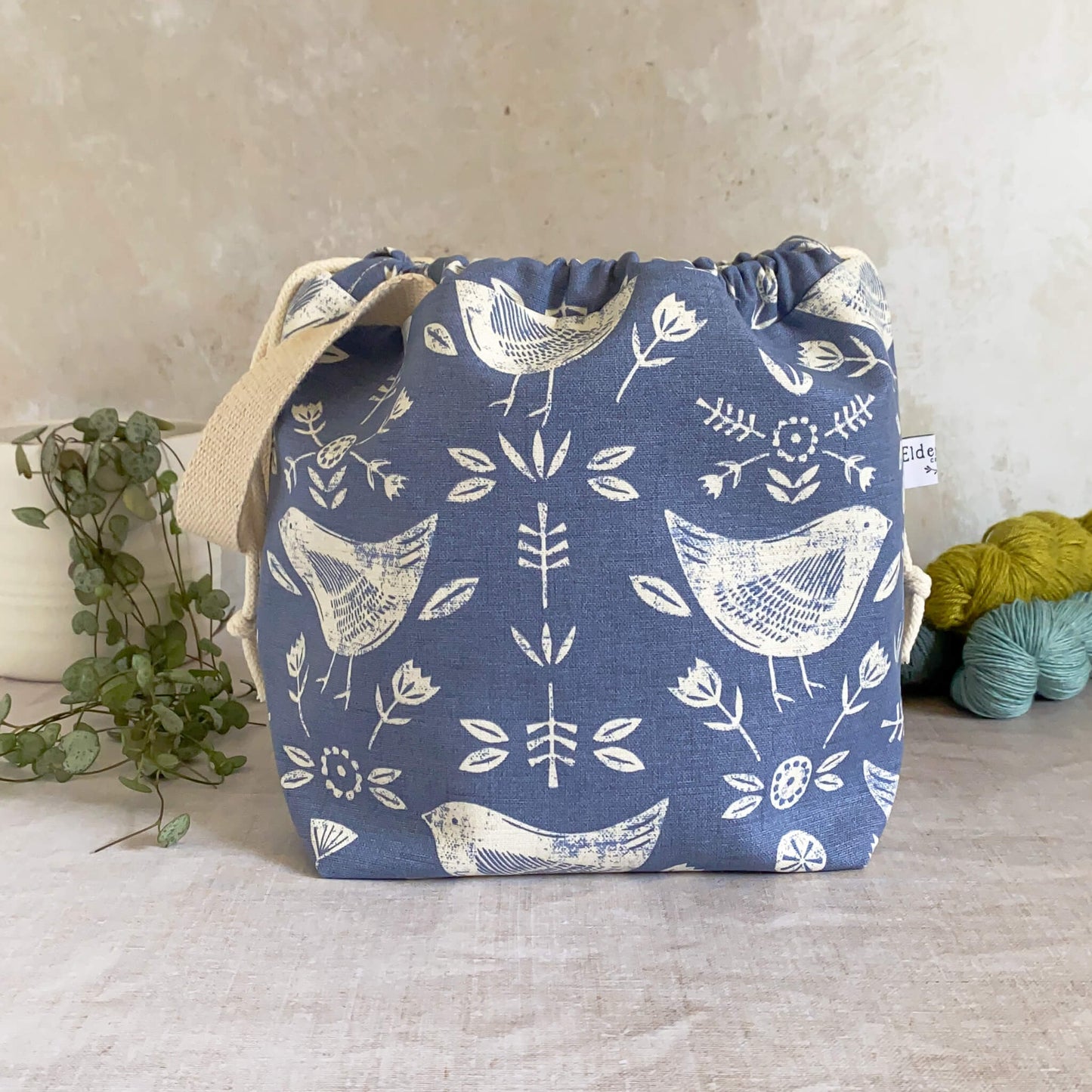 A medium-sized handmade project bag crafted from Scandi inspired blue bird-themed fabric, featuring a drawstring closure and sturdy handle, perfect for carrying knitting or crochet supplies. Designed for organizing shawl, scarf, and lightweight garment projects, its interior is lined with cream canvas and equipped with ample storage pockets. Displayed with three skeins of yarn and a potted plant.