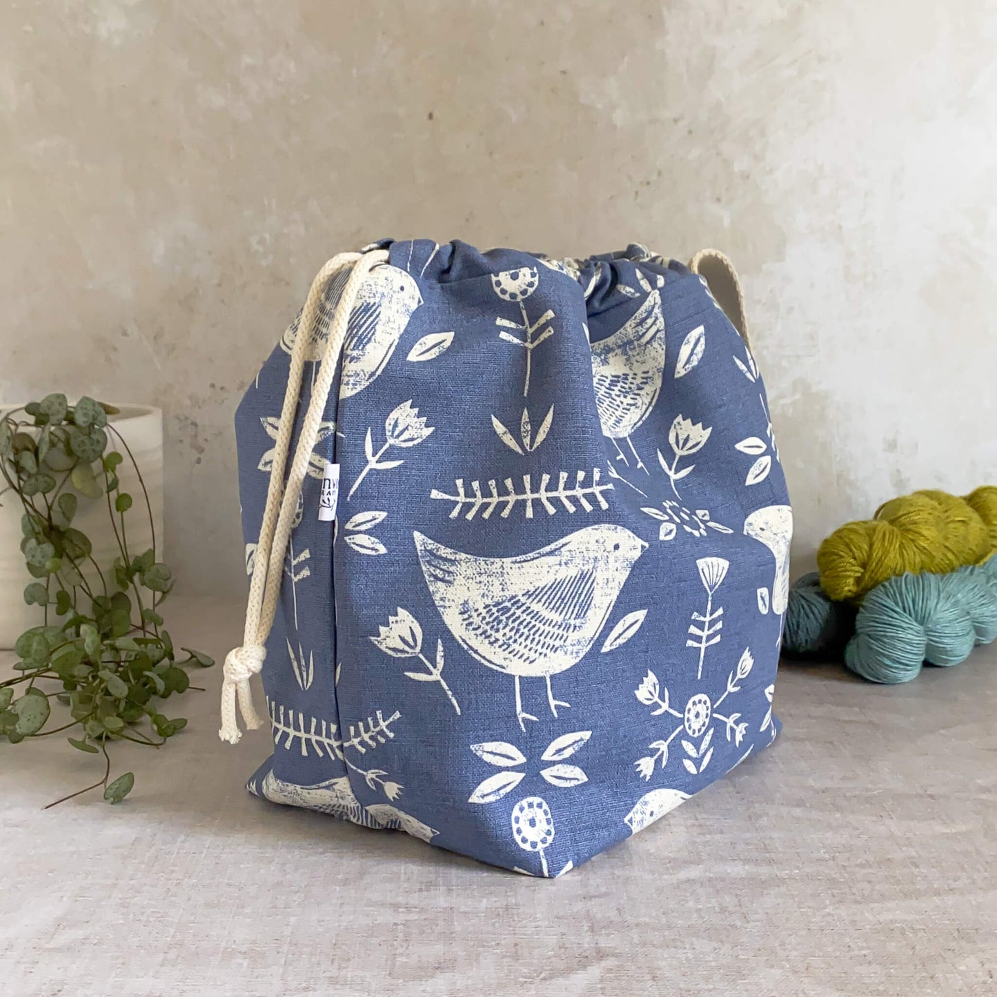 A side view of a medium-sized handmade project bag crafted from Scandi inspired blue bird-themed fabric, featuring a drawstring closure and sturdy handle. Ideal for organizing knitting or crochet supplies, it's tailored for shawl, scarf, and lightweight garment projects. The interior, lined with cream canvas, boasts numerous storage pockets. Displayed alongside three skeins of yarn and a potted plant.