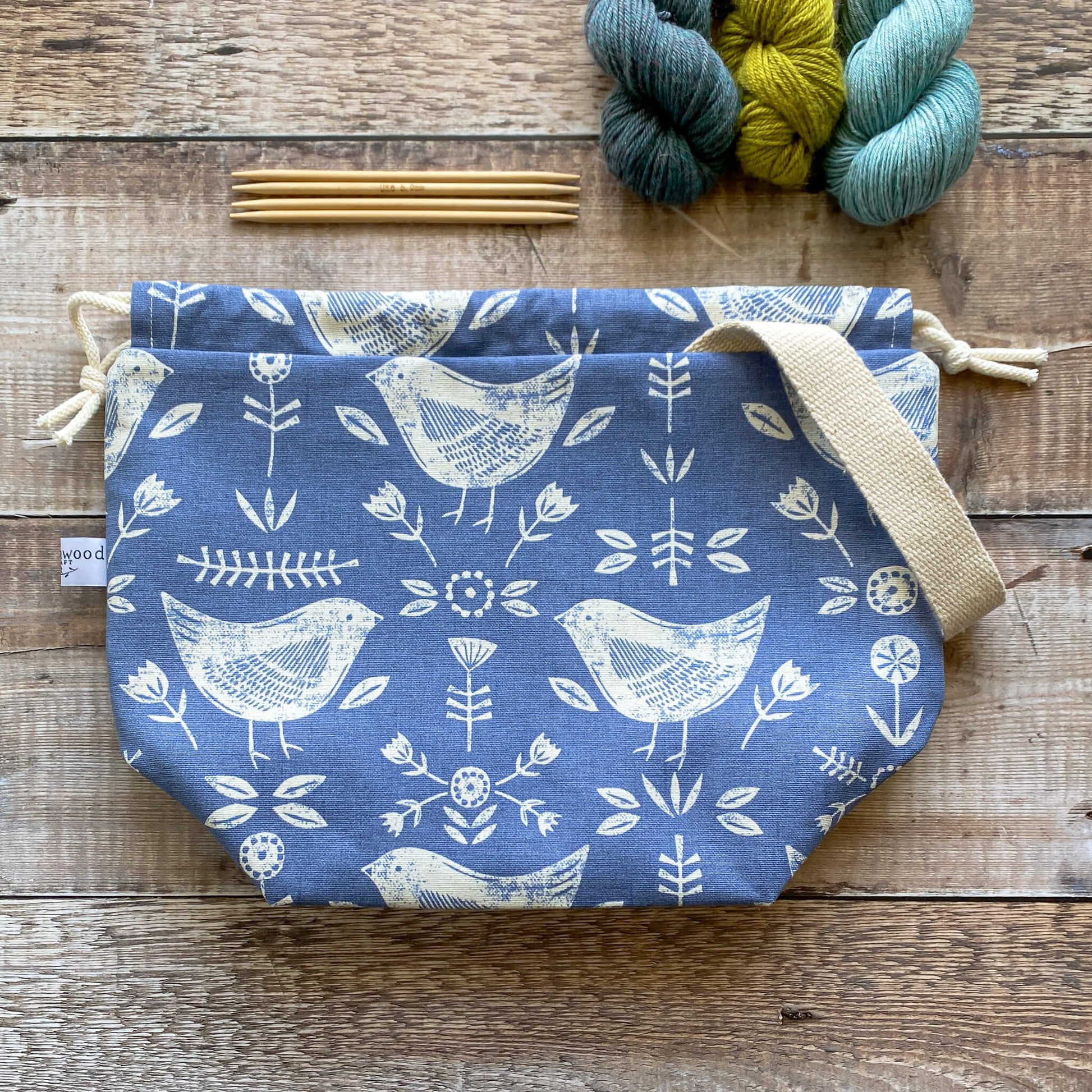 A medium sized handmade project bag in a blue Scandi inspired fabric, featuring a drawstring closure and sturdy handle, perfect for carrying knitting or crochet supplies. The bag is displayed next to three skeins of yarn and some wooden knitting needles. 