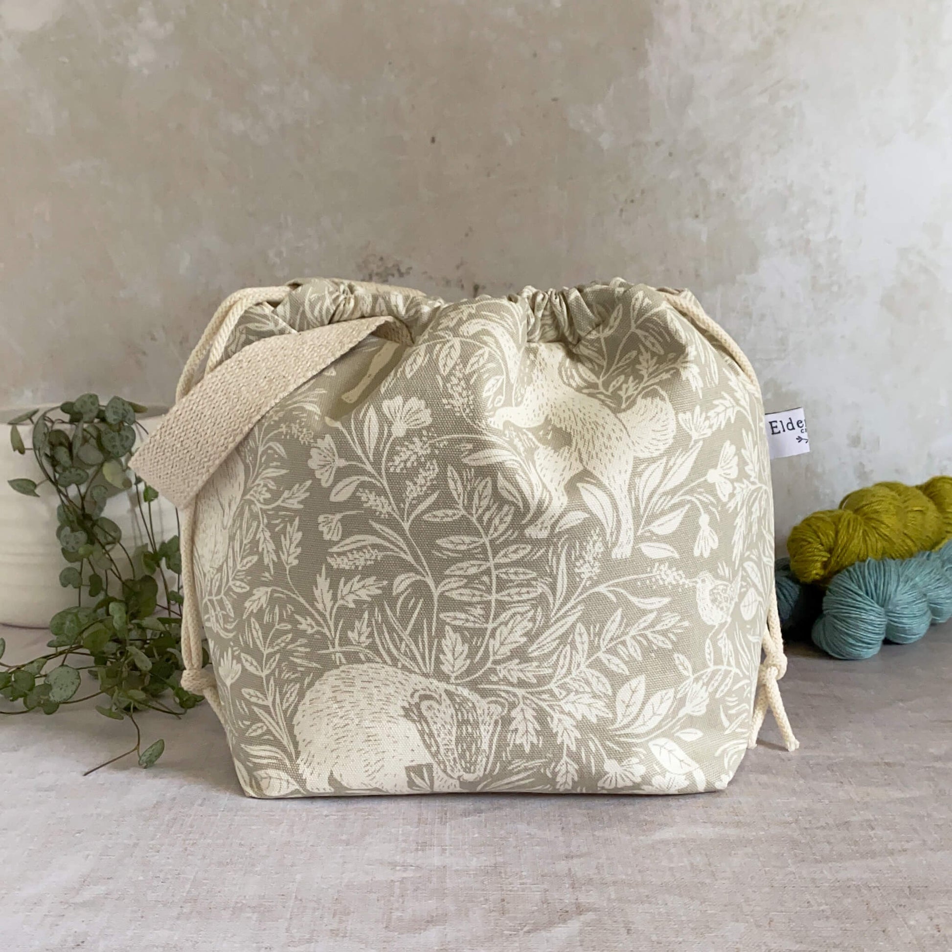 A medium sized handmade project bag in a linen coloured countryside themed fabric. A drawstring closure at the top, and a sturdy handle, ideal for carrying knitting or crochet supplies and projects. Ideal for organising shawl, scarf and lightweight garment projects. The interior of the bag is made from a cream canvas and has lots of storage pockets. The bag sits in front of three skeins of yarn and a pot plant. 