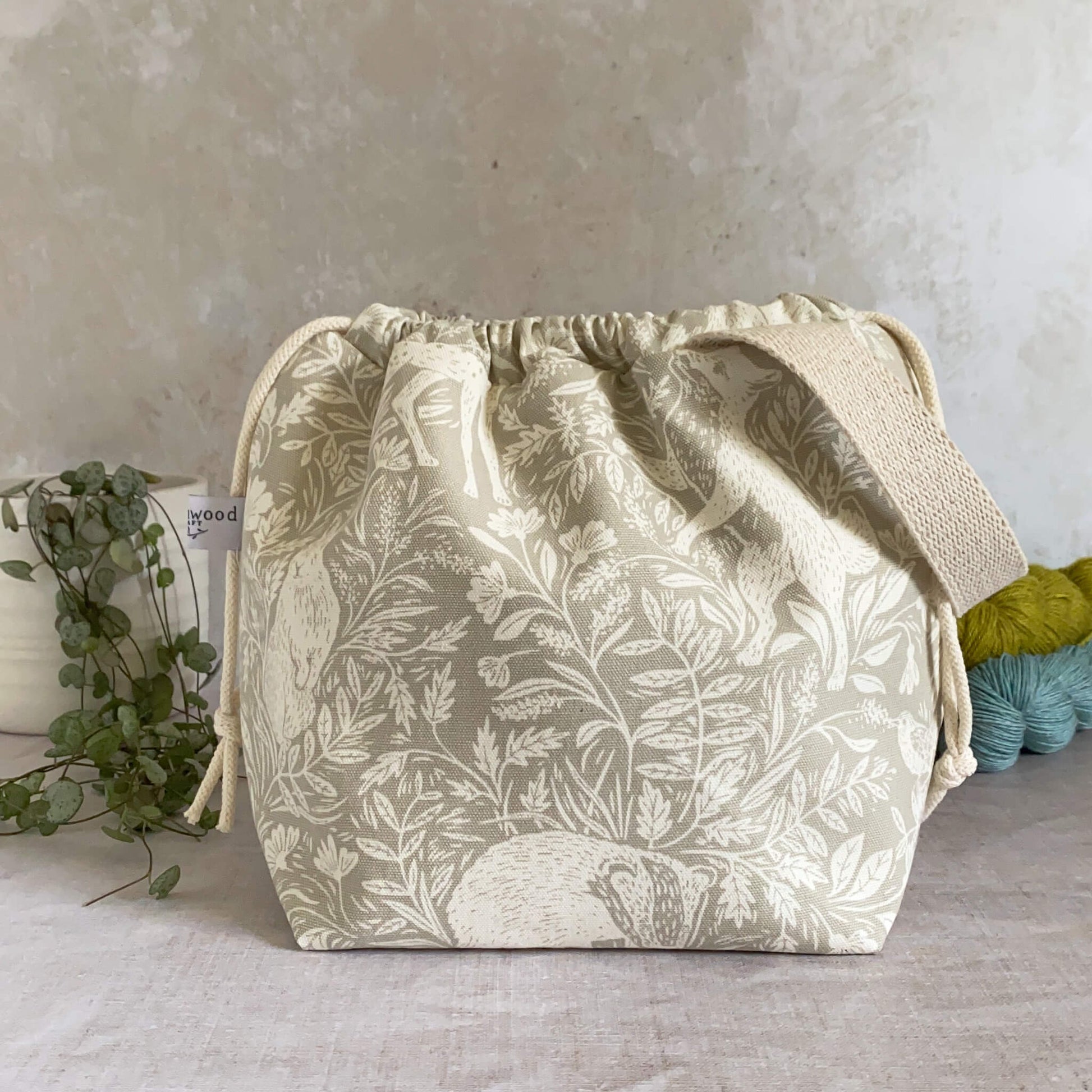 A medium-sized handmade project bag crafted from linen-colored countryside themed fabric, featuring a drawstring closure and sturdy handle, perfect for carrying knitting or crochet supplies. Ideal for organizing shawl, scarf, and lightweight garment projects. The interior, lined with cream canvas, offers ample storage pockets. Displayed alongside three skeins of yarn and a potted plant.