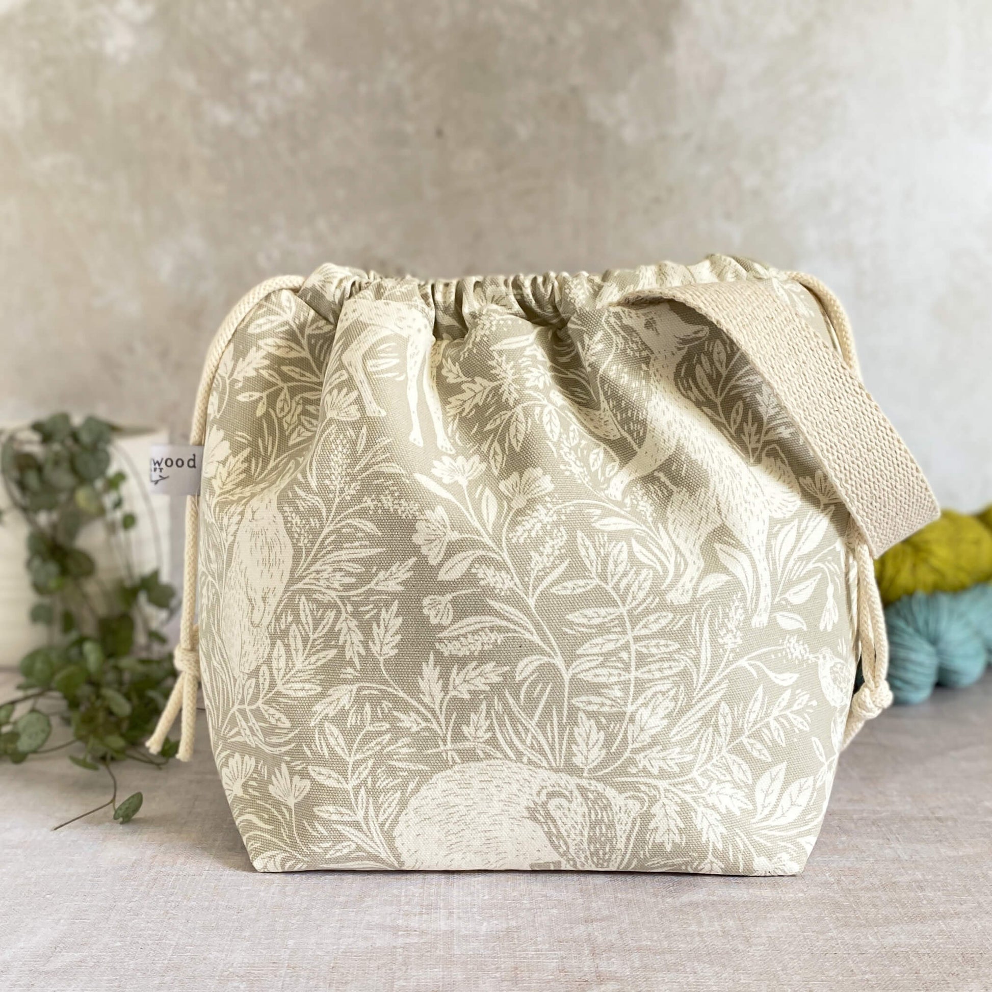 A medium-sized handmade project bag in linen-colored countryside themed fabric, featuring a convenient drawstring closure and sturdy handle, perfect for organizing knitting or crochet supplies. Ideal for storing shawl, scarf, and lightweight garment projects, its interior is lined with cream canvas and equipped with numerous storage pockets. Displayed alongside three skeins of yarn and a potted plant.