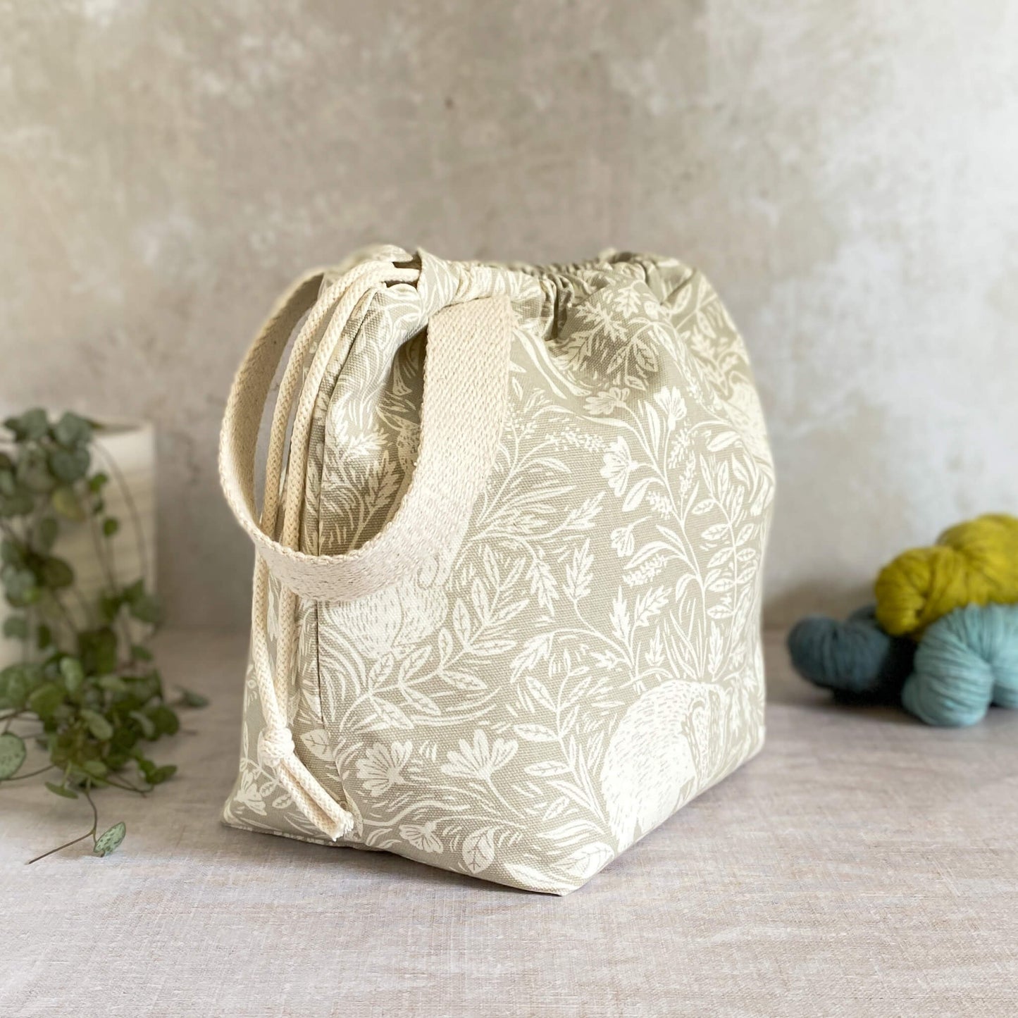 A side view of a medium-sized handmade project bag crafted from linen-colored countryside themed fabric, featuring a practical drawstring closure and sturdy handle. Ideal for organizing knitting or crochet supplies, it's designed for storing shawl, scarf, and lightweight garment projects. The interior is lined with cream canvas and includes multiple storage pockets, displayed alongside three skeins of yarn and a potted plant.