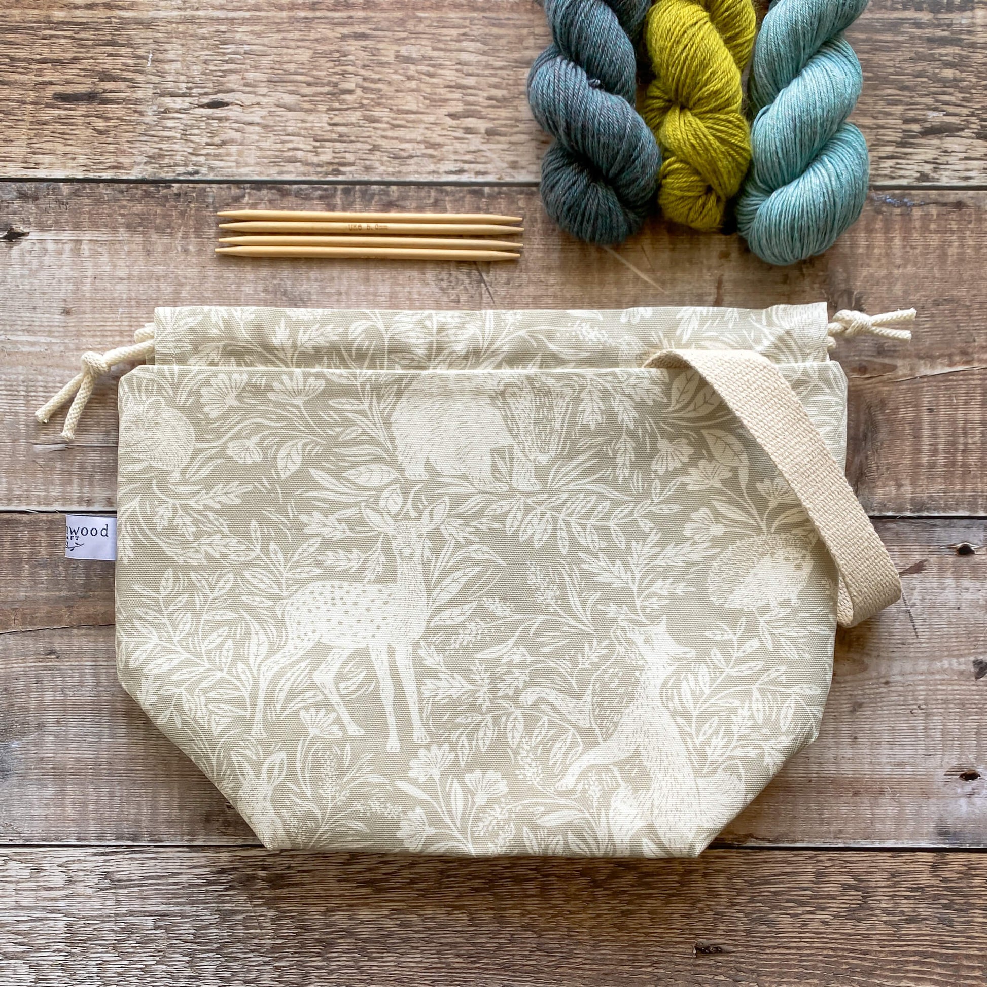 A medium sized handmade project bag in a linen coloured countryside fabric, featuring a drawstring closure and sturdy handle, perfect for carrying knitting or crochet supplies. The bag is displayed next to three skeins of yarn and some wooden knitting needles. 