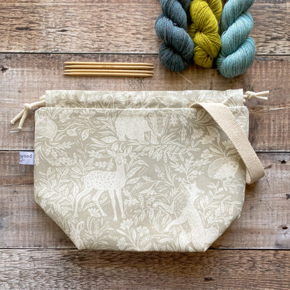 A medium sized handmade project bag in a linen coloured countryside fabric, featuring a drawstring closure and sturdy handle, perfect for carrying knitting or crochet supplies. The bag is displayed next to three skeins of yarn and some wooden knitting needles. 