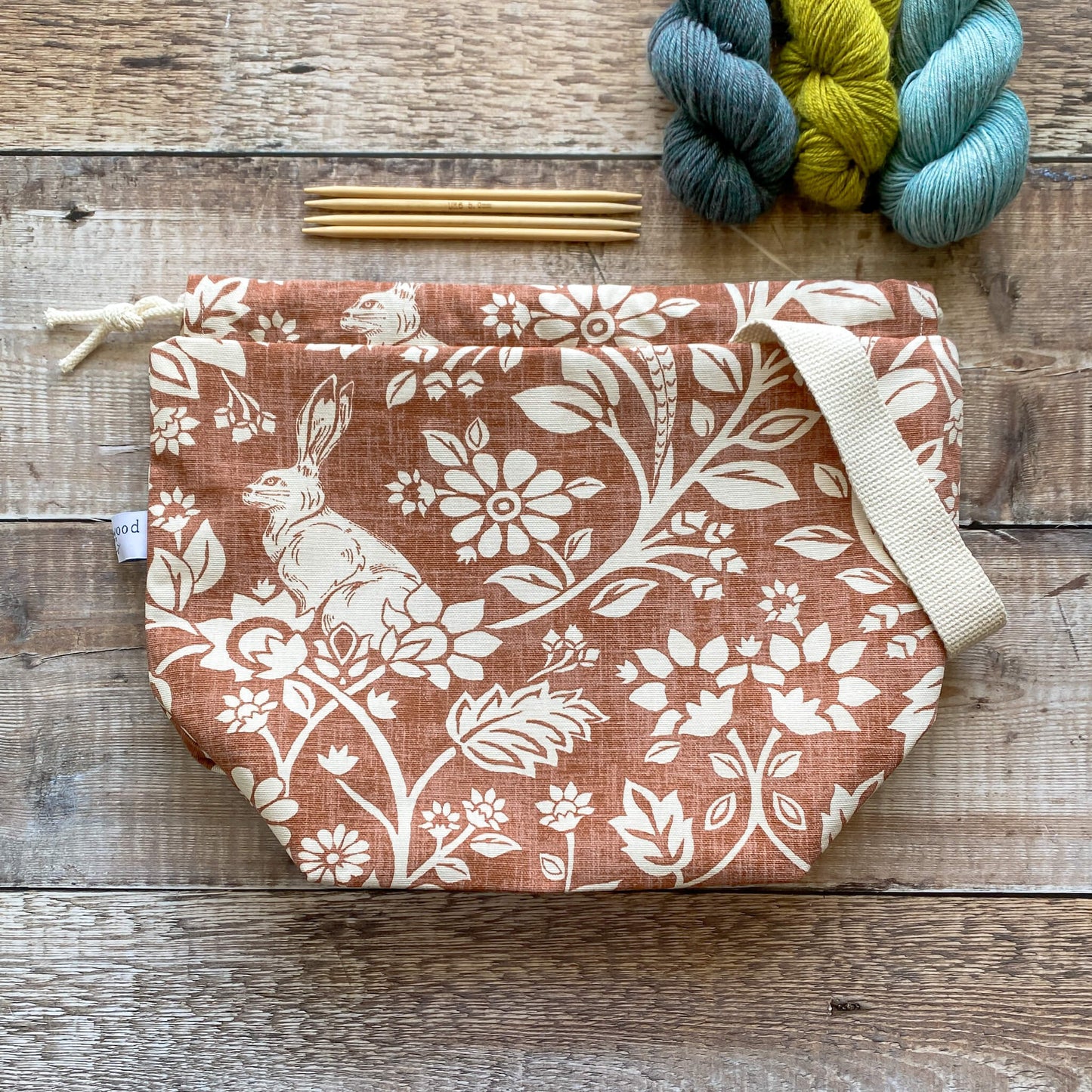 A medium handmade project bag in heathland themed fabric, designed with a drawstring closure and sturdy handle, ideal for carrying knitting or crochet supplies. Displayed alongside three skeins of yarn and wooden knitting needles.