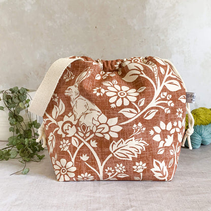 A medium-sized handmade project bag crafted from heathland themed fabric, featuring a convenient drawstring closure and sturdy handle, perfect for organizing knitting or crochet supplies and ideal for projects such as shawls, scarves and smaller garments. The interior is lined with cream canvas and includes multiple storage pockets. Displayed alongside three skeins of yarn and a potted plant.