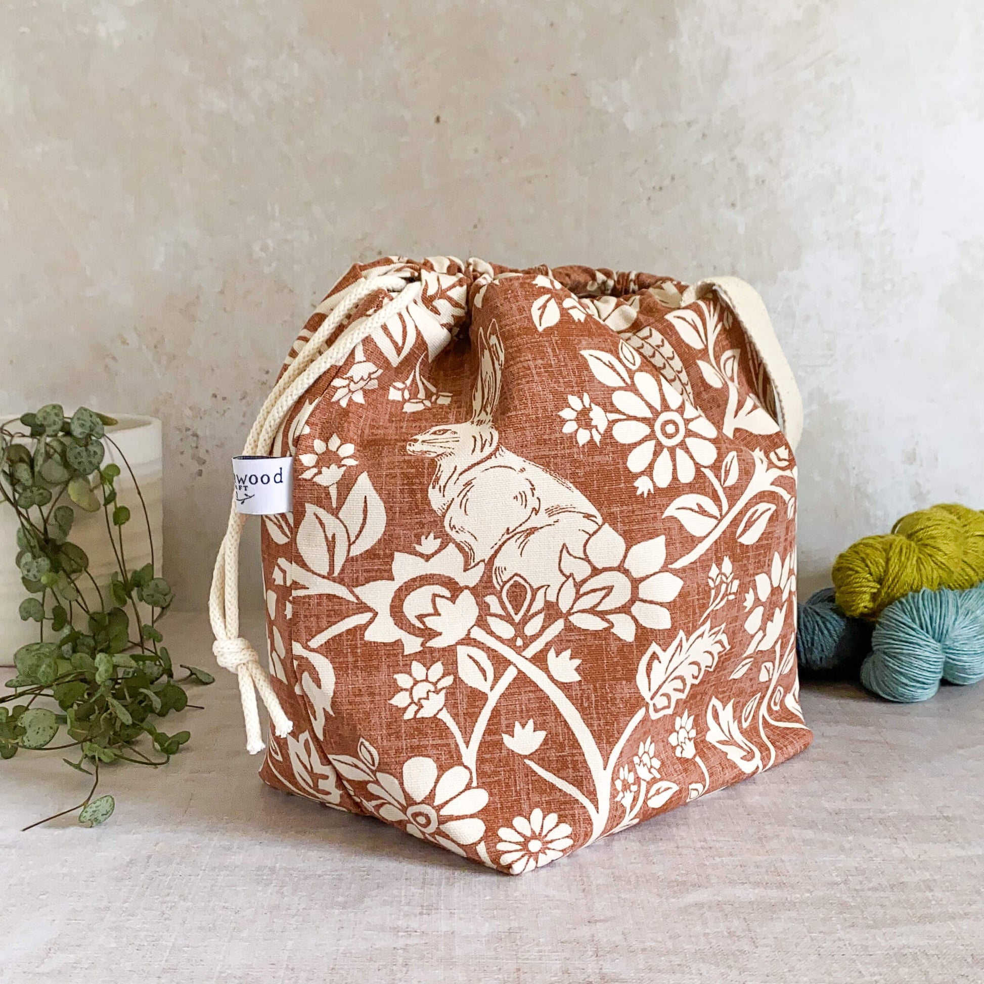 A side view of a medium-sized handmade project bag crafted from heathland themed fabric, showcasing its convenient drawstring closure and sturdy handle. Ideal for organizing knitting or crochet supplies, it's well-suited for projects such as shawls, scarves, and smaller garments. The interior is lined with cream canvas and features multiple storage pockets. Displayed next to three skeins of yarn and a potted plant.