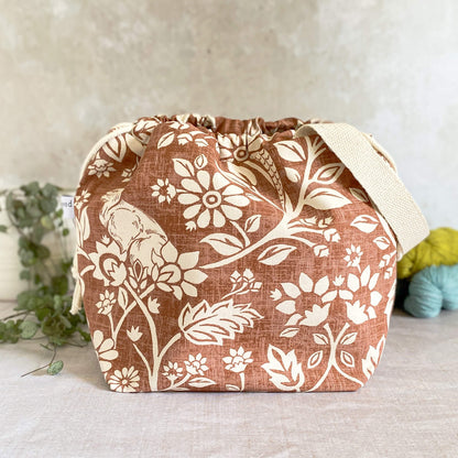 A medium-sized handmade project bag made from heathland themed fabric, with a practical drawstring closure and sturdy handle, ideal for neatly organizing knitting or crochet supplies. Its interior is lined with cream canvas and boasts multiple storage pockets. Displayed next to three skeins of yarn and a potted plant.