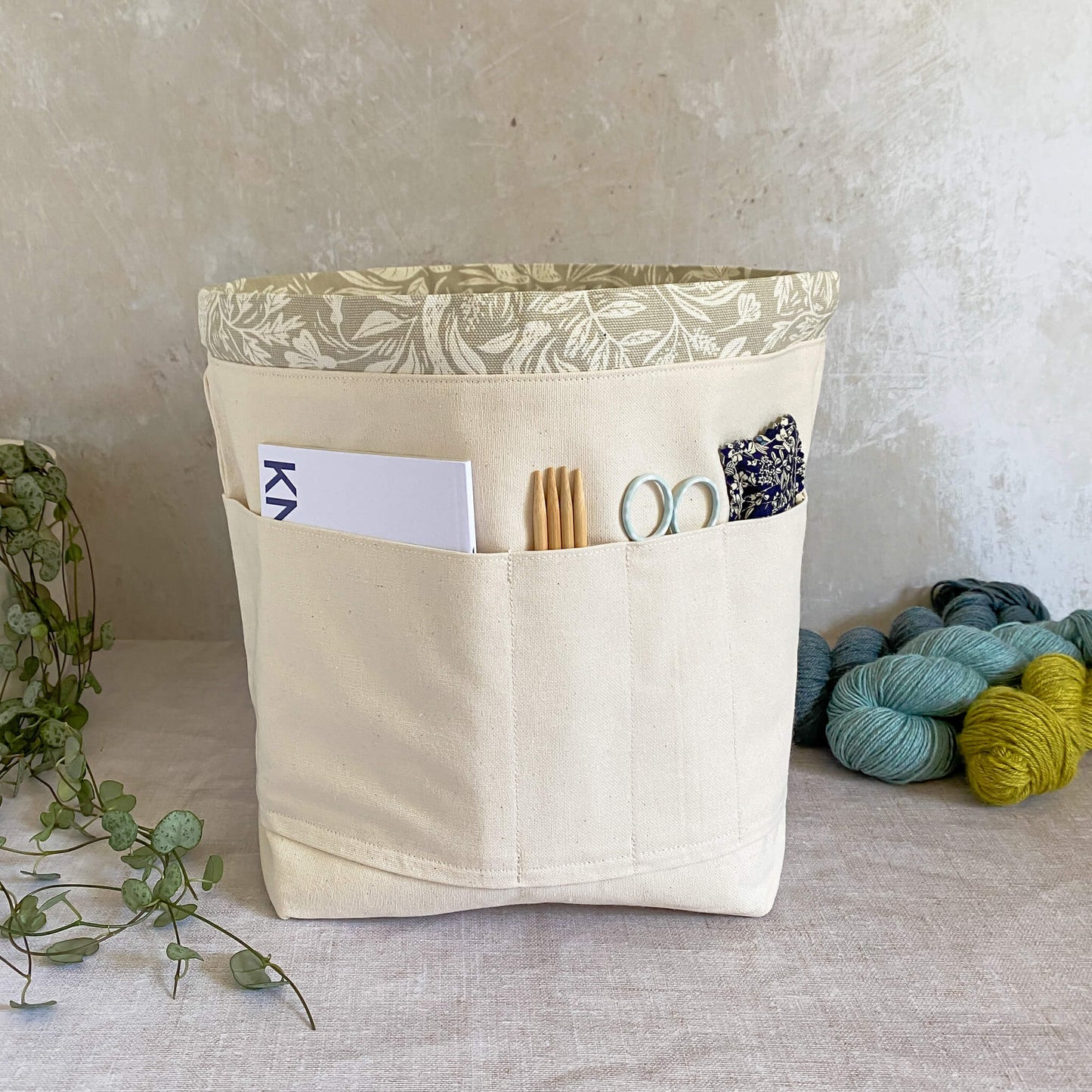 Interior view of a knitting and crochet project bag, showcasing pockets on one side for holding knitting or crochet patterns, a notebook, pen, needles, hooks, and other crafting accessories. The interior features durable natural-colored canvas