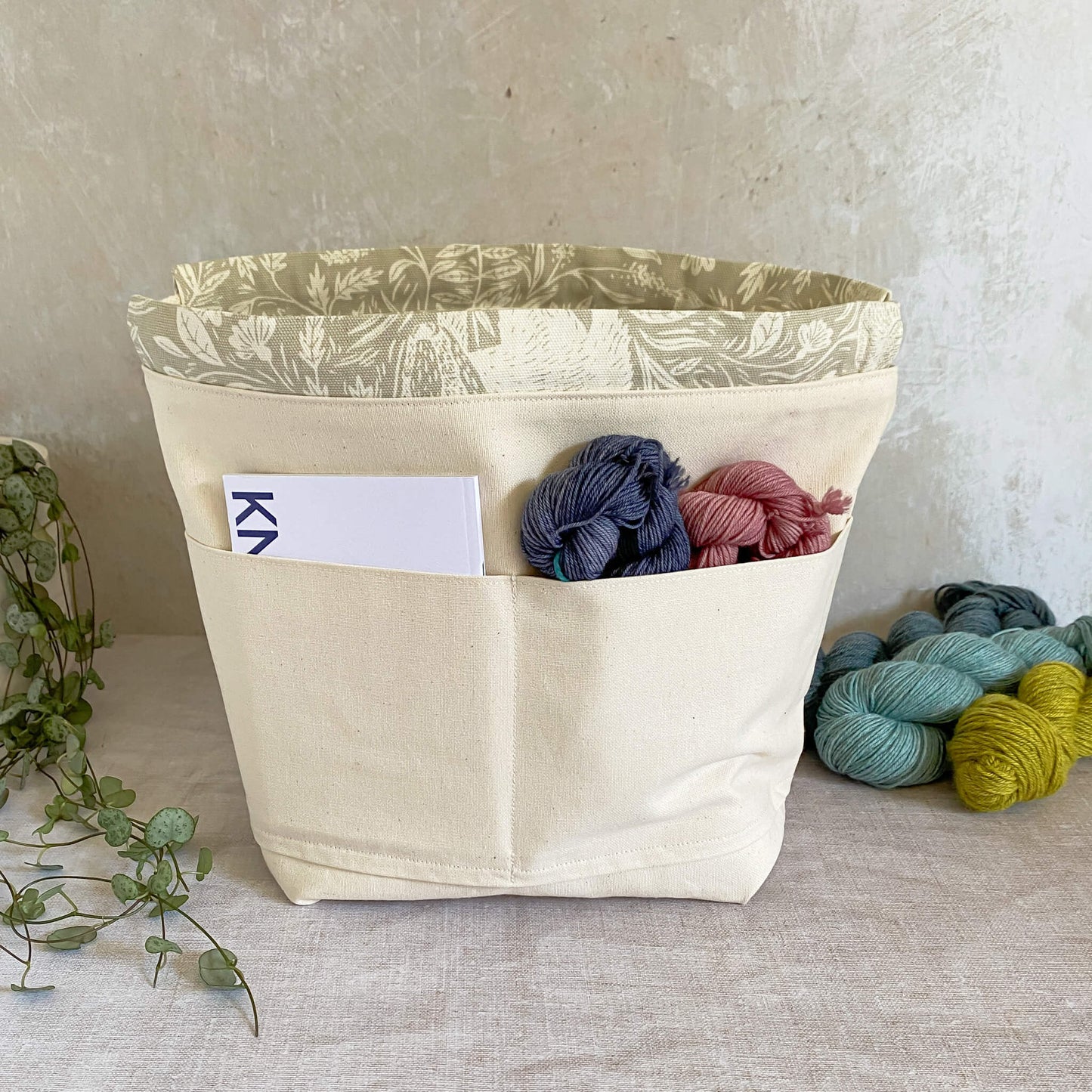 Interior view of a knitting and crochet project bag, highlighting pockets on one side for storing knitting or crochet patterns, a notebook, pen, needles, hooks, and other crafting accessories. The interior is lined with durable natural-colored canvas.