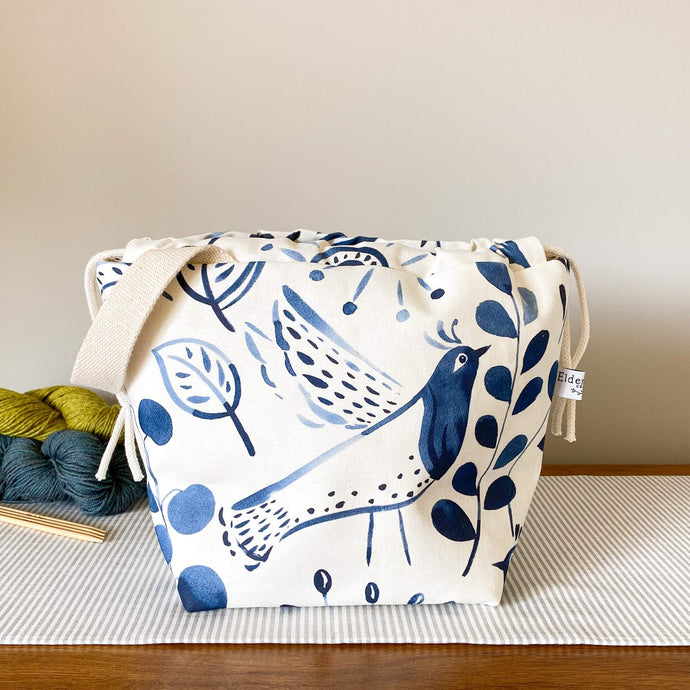 Image alt text: A knitting project bag, adorned with a folk art indigo-colored print, rests on a table top. Three skeins of yarn are arranged neatly behind the bag, creating a colorful backdrop.