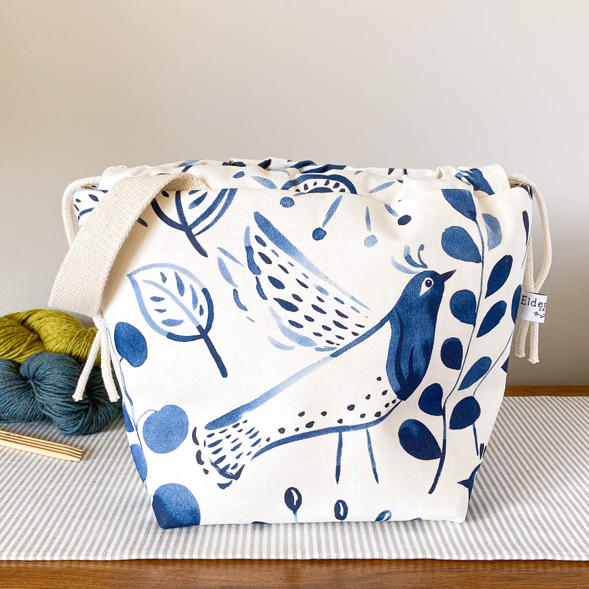 Alt text: A knitting project bag made of fabric with a folk art indigo print sits on a table. Three colorful skeins of yarn are positioned behind the bag, adding a vibrant touch.