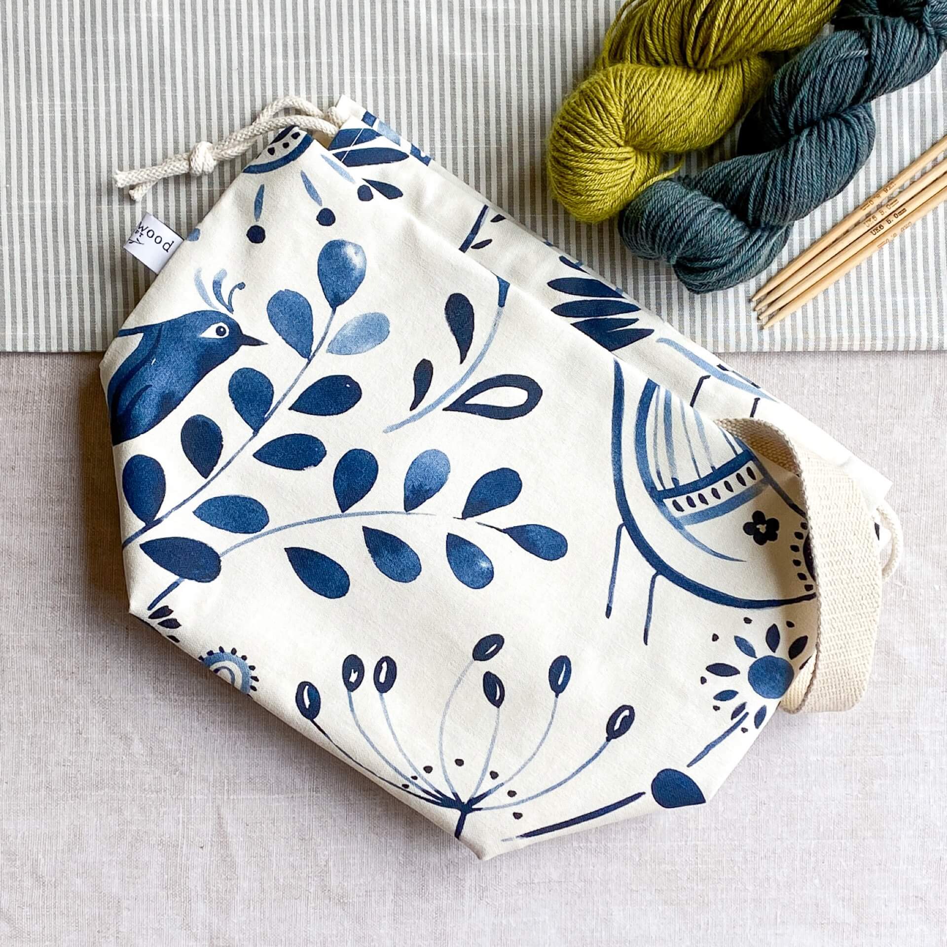 An indigo-colored folk art print knitting project bag placed on a table. Above the bag, two colorful skeins of yarn are neatly arranged next to wooden knitting needles. The bag features a vibrant and intricate folk art design on its fabric.