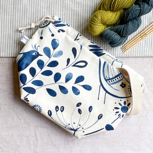 An indigo-colored folk art print knitting project bag placed on a table. Above the bag, two colorful skeins of yarn are neatly arranged next to wooden knitting needles. The bag features a vibrant and intricate folk art design on its fabric.