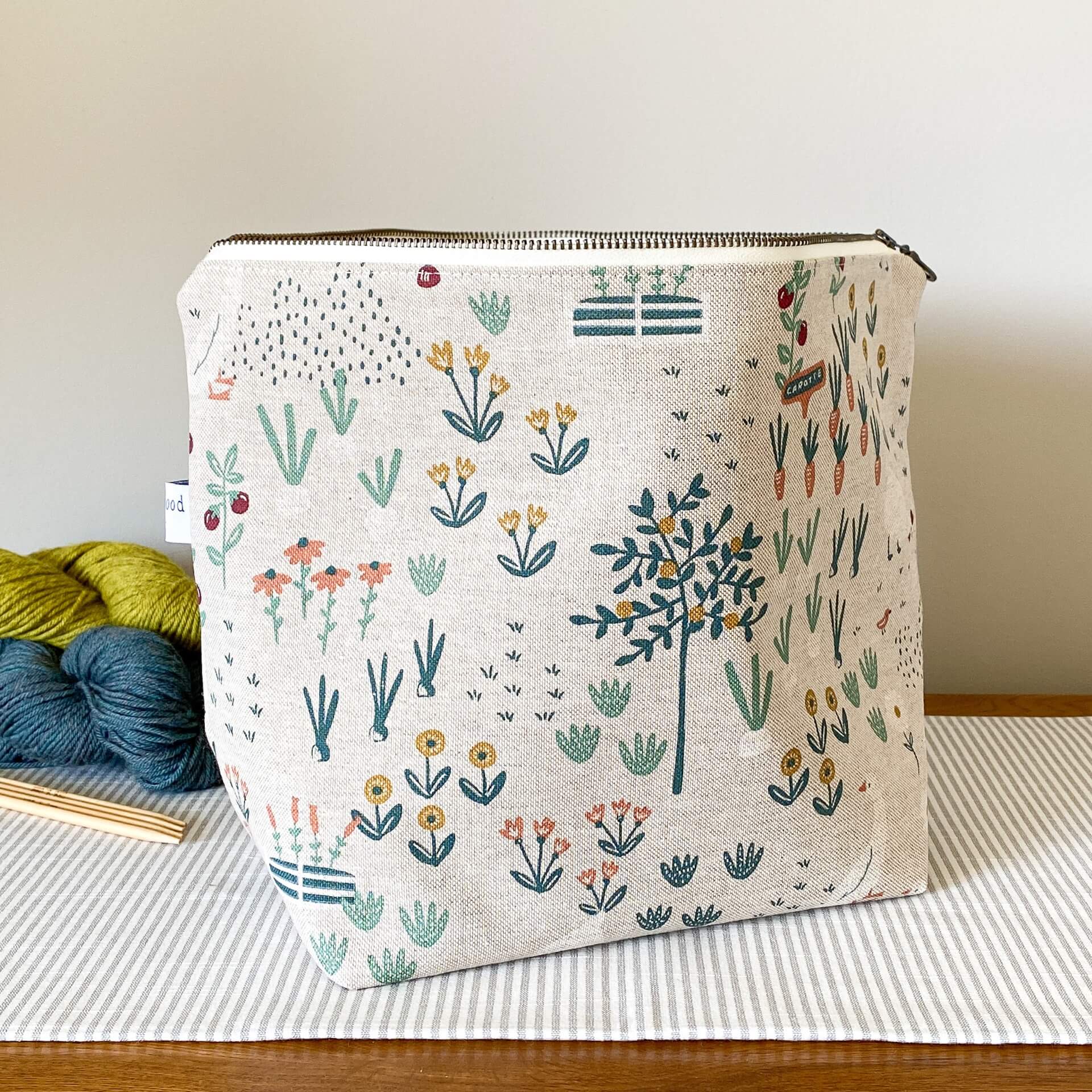 A zippered project bag sits on a table top. Behind the bag are some skeins of yarn in different colours neatly arranged. Also in view are some wooden knitting needles. The bag is at the centre of the image and is made from a fabric that depicts a cottage garden. 