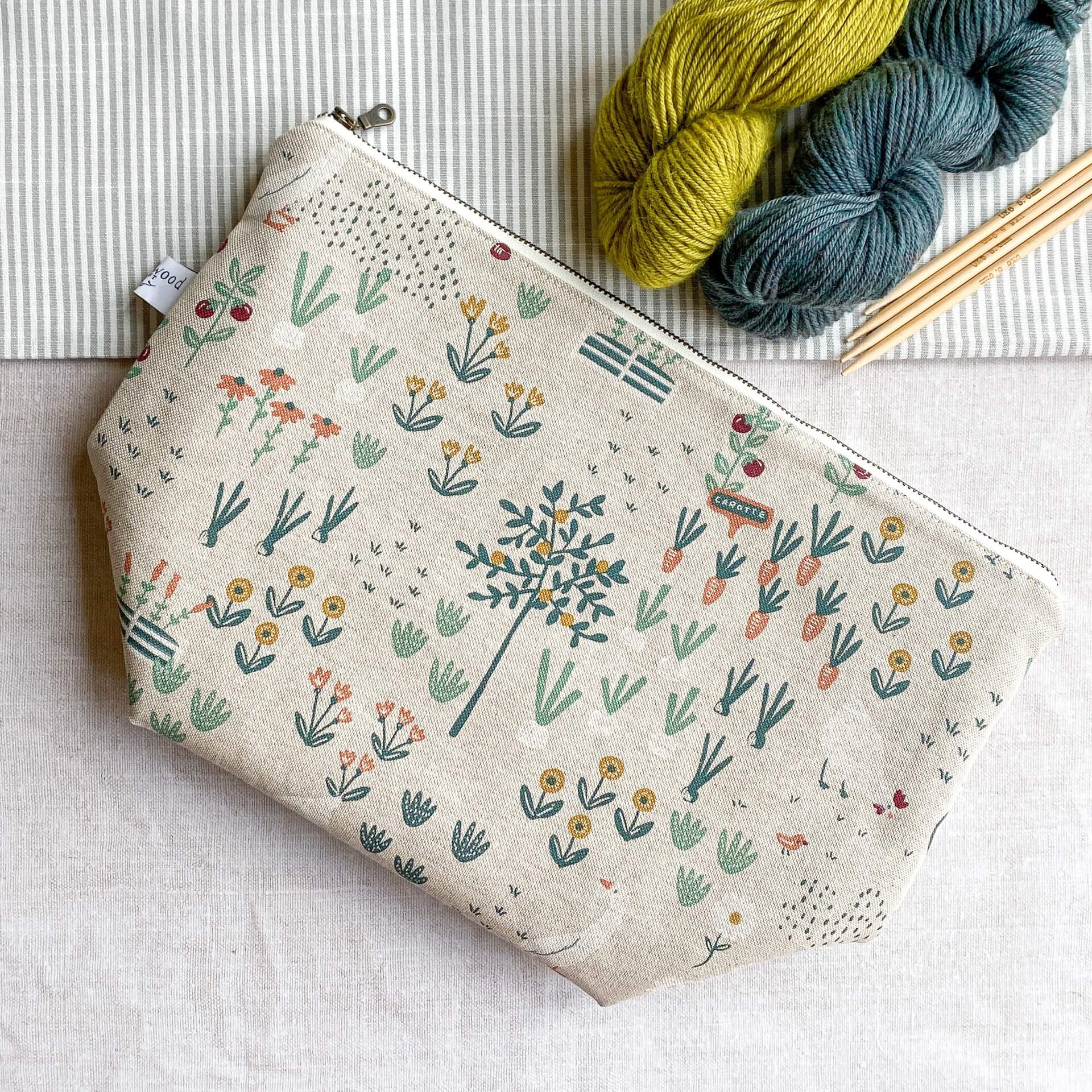 A zippered project bag for knitting lies on a linen covered table top next to two skeins of yarn - one green and one blue - and some wooden knitting needles. 