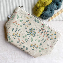 Load image into Gallery viewer, A zippered project bag for knitting lies on a linen covered table top next to two skeins of yarn - one green and one blue - and some wooden knitting needles. 
