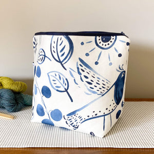 A knitting project bag with folk art design in indigo print rests on a table. Three colorful skeins of yarn are placed behind the bag, creating a vibrant backdrop.