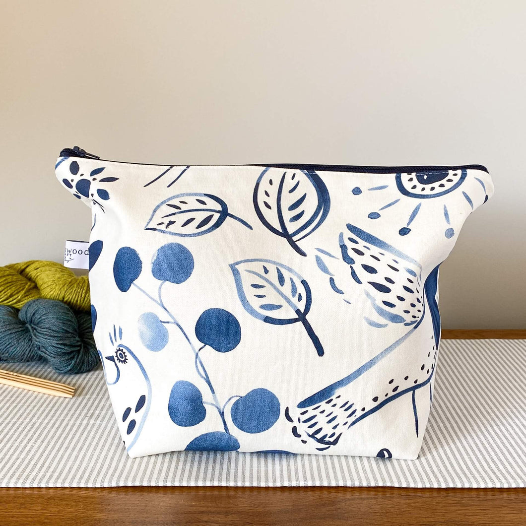 An indigo-colored knitting project bag sits on a table top. The bag features a folk art print fabric, depicting intricate patterns and motifs. Behind the bag, three skeins of yarn in different colors are neatly arranged.