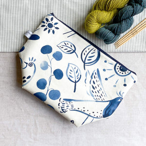  A knitting project bag placed on a table. The bag is made of fabric with a folk art image printed in indigo color. It features various motifs like leaves, birds, and flowers.