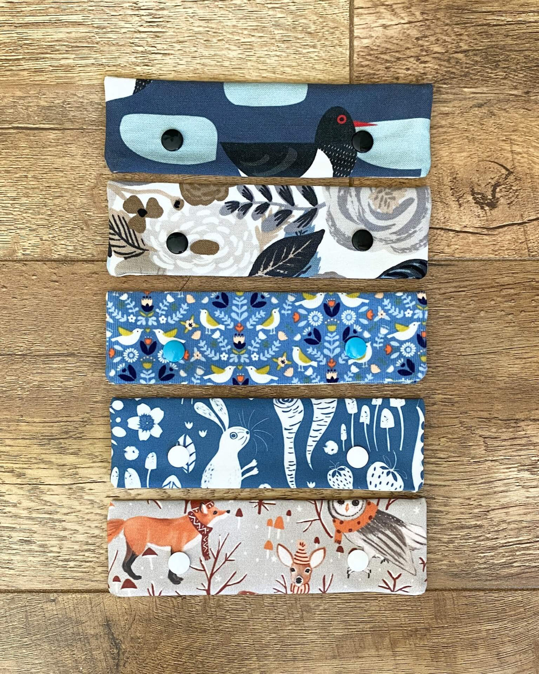 A line of five DPN holders for knitting projects are on a wooden floor. They are made from a variety of different fabrics all featuring something from the natural world such as animals, birds or flowers. 