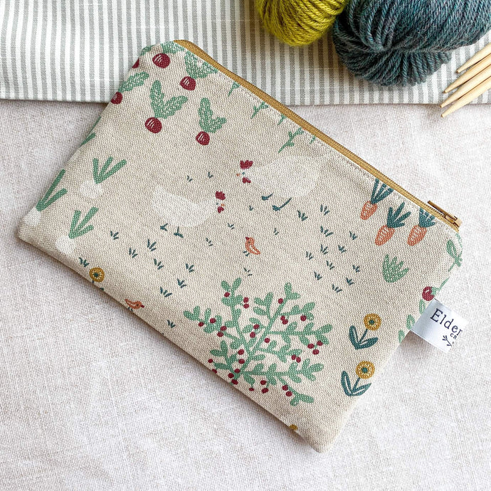 A small zipped pouch intended to be used for knitting notions lies on a table top next to two skeins of yarn and some wooden knitting needles. On the front of the pouch we can see some chickens, a bird, some beetroot and carrots, all printed in a whimsical style. 